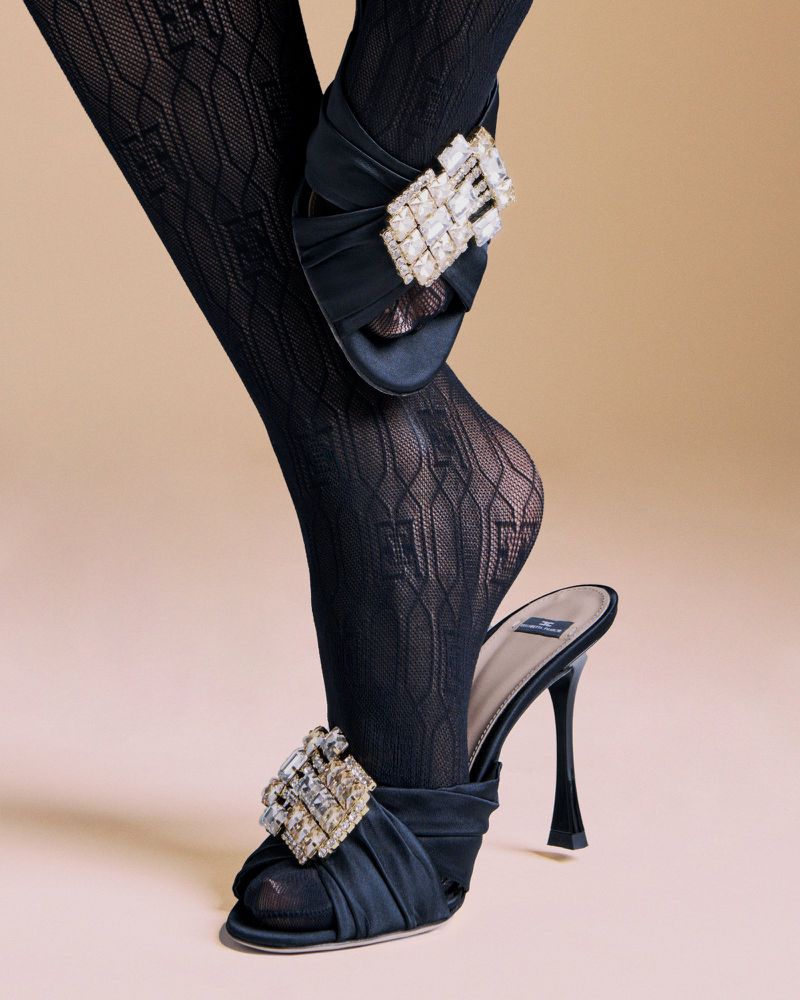 Close-up studio shot of woman's legs wearing black patterned stockings and black high-heeled mules with crystal embellishments.