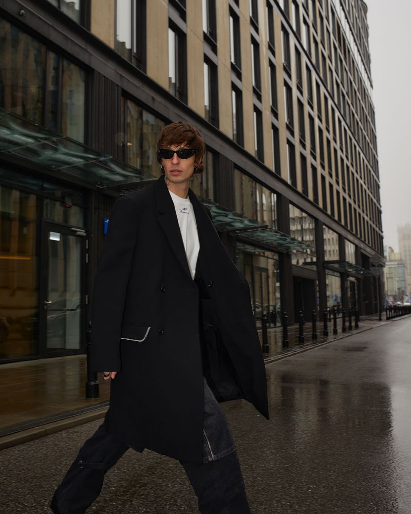 A male fashion model poses in a black coat and dark jeans on a city street with modern architecture and wet pavement.
