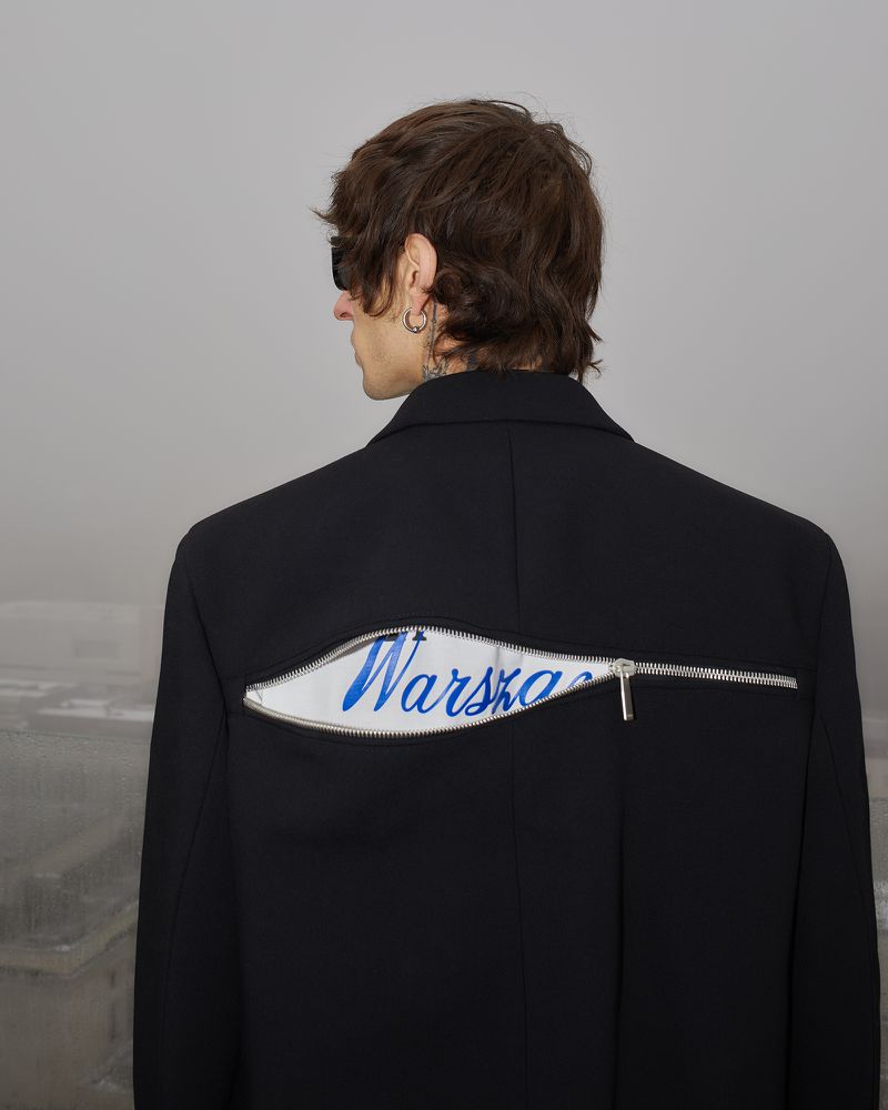 Man wearing a black blazer with a zipper design, photographed from the back, showcasing fashion in an outdoor urban setting.