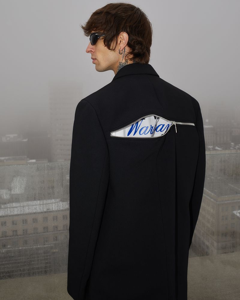 Adult male in black jacket with 'Waray' text design stands against an overcast cityscape, captured in a medium shot highlighting fashion details.