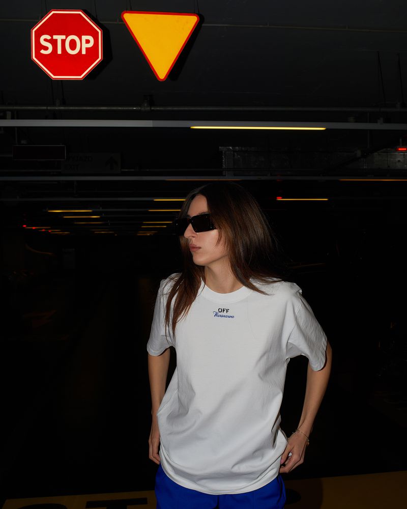 Stylish woman in sunglasses and casual clothing stands in a dark parking garage, with overhead traffic signs creating a unique composition.