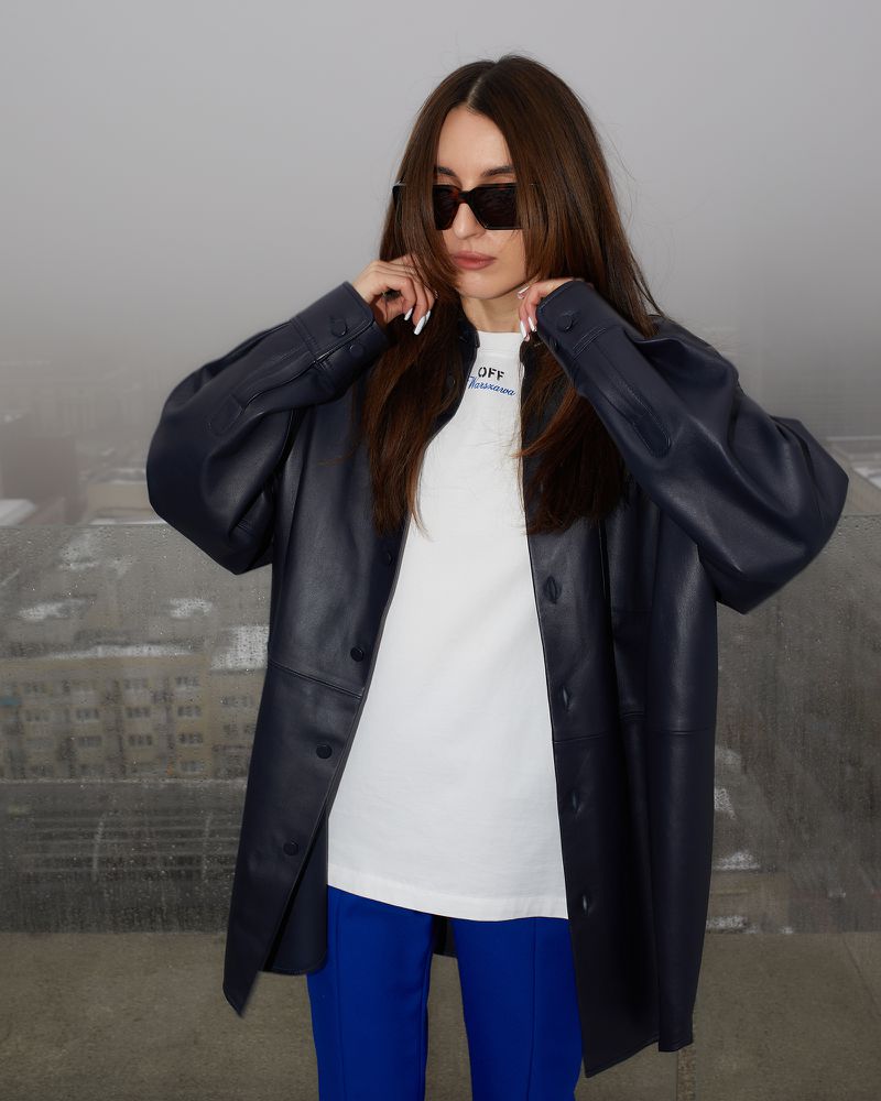 Stylish adult woman with sunglasses and a blue jacket posing outdoors against a blurred urban backdrop in a medium studio shot.
