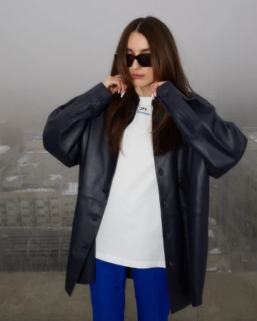 Stylish adult woman with sunglasses and a blue jacket posing outdoors against a blurred urban backdrop in a medium studio shot.