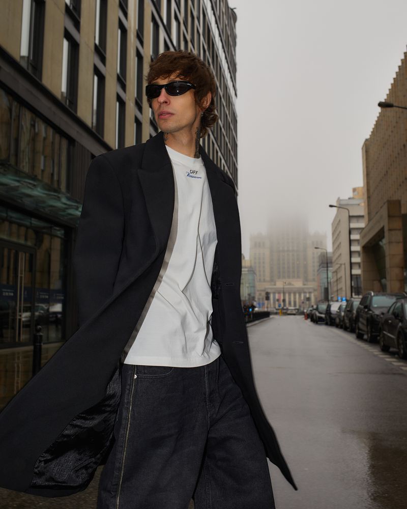 Adult male poses in an urban street wearing dark sunglasses, a white t-shirt, dark jeans, and a black coat with modern buildings behind him.