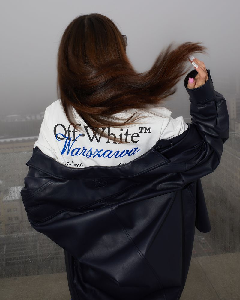 Adult female with long brown hair standing in a dark jacket and Off-White hoodie near a glass railing overlooking a foggy cityscape.