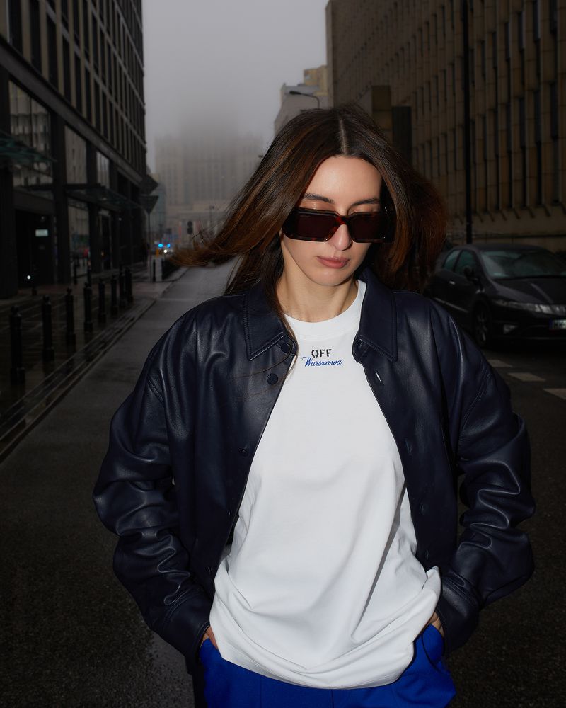 Woman with long brown hair in a navy jacket, white shirt, and blue pants stands on a city street with tall buildings.