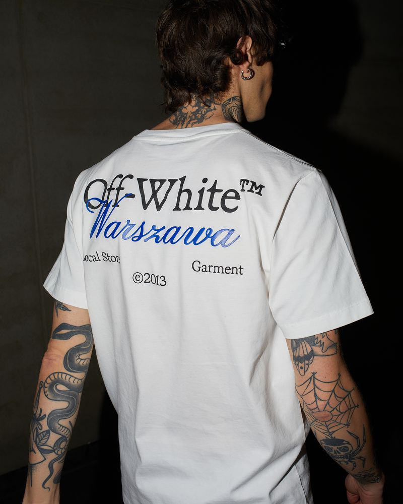 Back view of an adult male wearing a white Off-White T-shirt with blue 'Warszawa' text, featuring snake and spiderweb tattoos on his arms.
