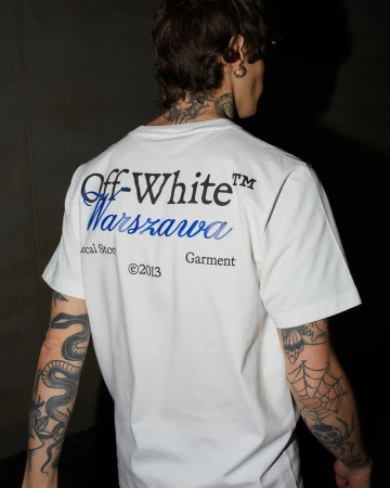 Back view of an adult male wearing a white Off-White T-shirt with blue 'Warszawa' text, featuring snake and spiderweb tattoos on his arms.