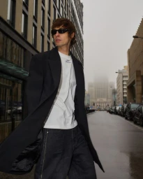 Man in dark sunglasses, a long black overcoat, and a white t-shirt standing on a wet city street in front of urban buildings.