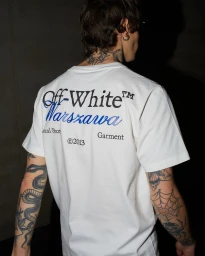 Back view of a man with arm tattoos wearing a white Off-White Warszawa graphic t-shirt against a dark background.