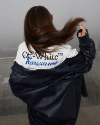 Woman with long brown hair wearing a dark jacket over a white Off-White Warszawa sweatshirt