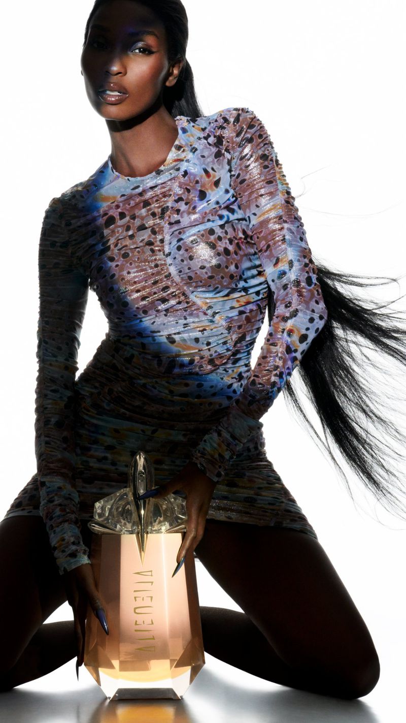 Elegant dark-skinned woman in patterned mini dress poses kneeling next to a perfume bottle in a bright studio setting.