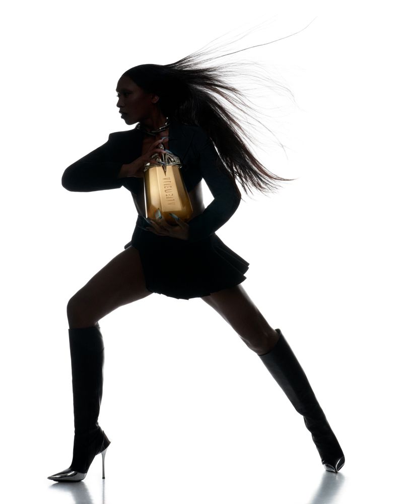 Silhouette of a Black female fashion model in motion, wearing a skirt suit and boots, holding a gold perfume bottle against white.
