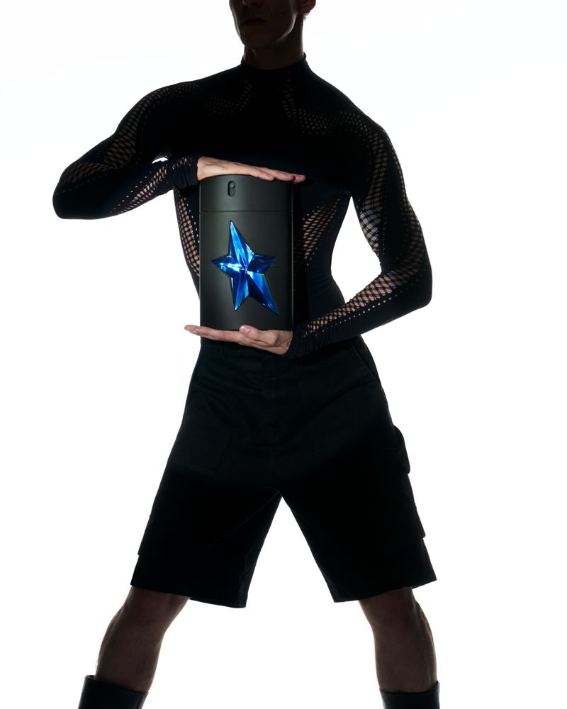 Black cylindrical bottle with blue star logo in the hands of a slim adult male in a black mesh shirt, silhouetted against a white background.