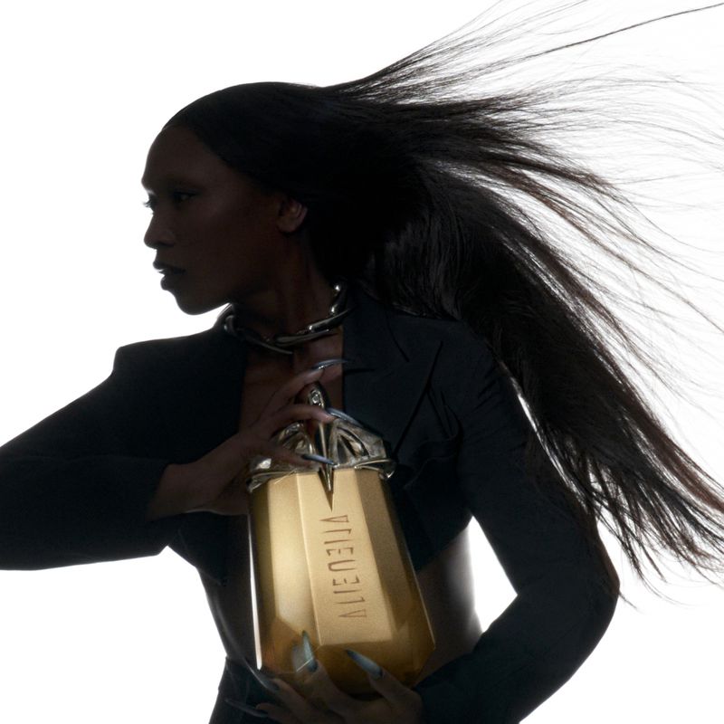 Elegant Black woman with flowing hair holds gold Alien Goddess perfume bottle in a beauty shot against a white background, studio lighting.