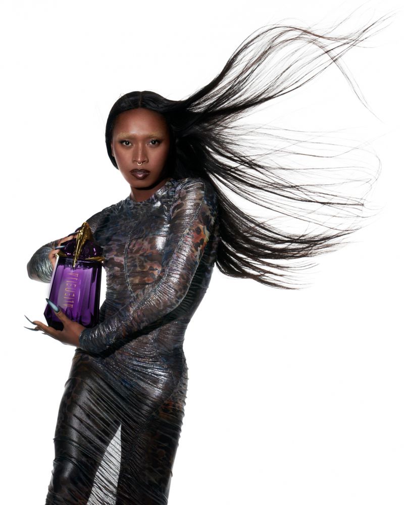 Woman in snakeskin-pattern dress holding a purple geometric-shaped perfume bottle in a white studio with hair blowing in the wind.