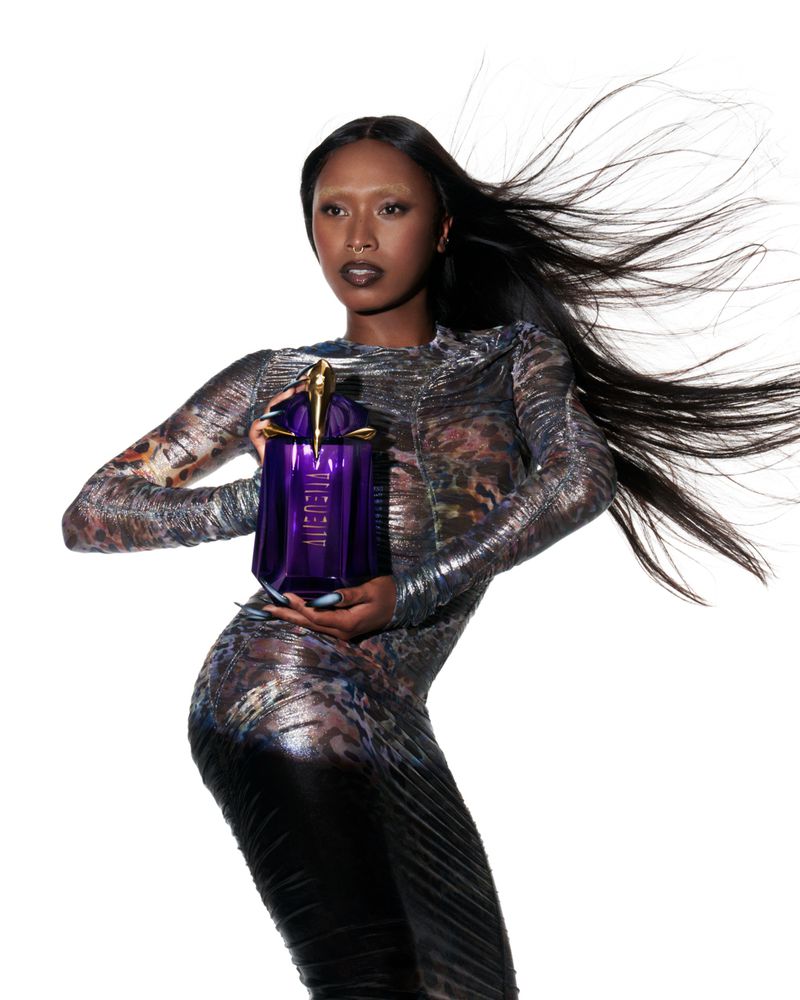 Woman with long, dark hair holds a purple Alien Goddess perfume bottle against a stark white background in a beauty shot.