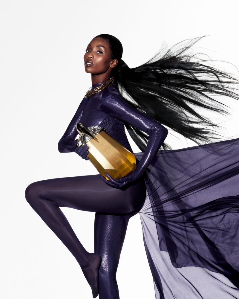 Fashion shot of a Black female model in a purple bodysuit, holding a gold perfume bottle against a white background. Ponytail hairstyle.