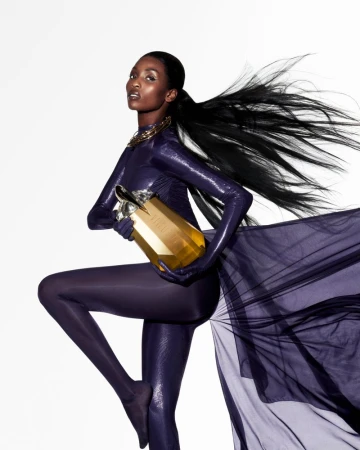 Model in purple catsuit holding a gold fragrance bottle against a clean white background
