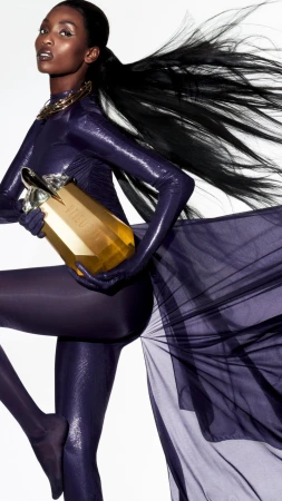 Model in shiny purple catsuit holding a large gold perfume bottle against a white background