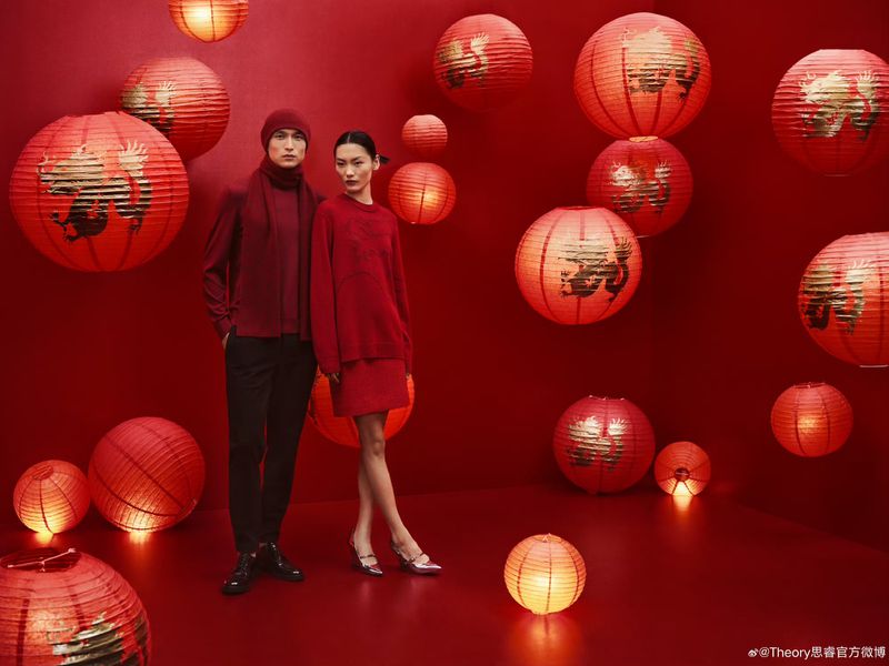 Two fashion models in red attire pose amidst a backdrop of festive red Chinese lanterns, creating a vibrant, celebratory scene.