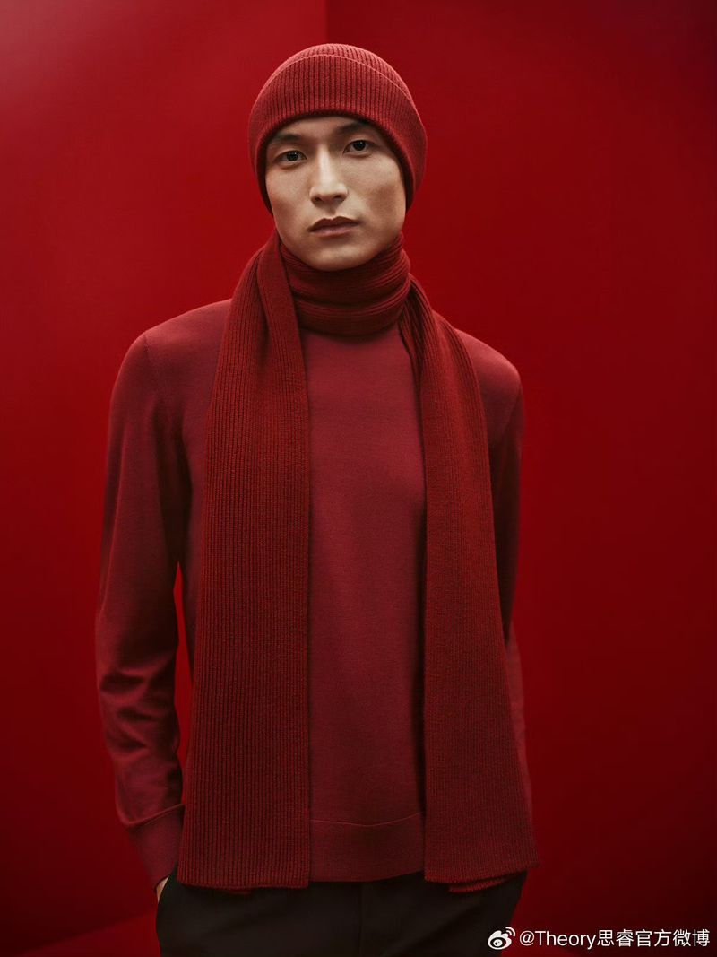 Portrait of a male model wearing a red knit beanie, scarf, and sweater against a monochromatic red backdrop in a studio setting.