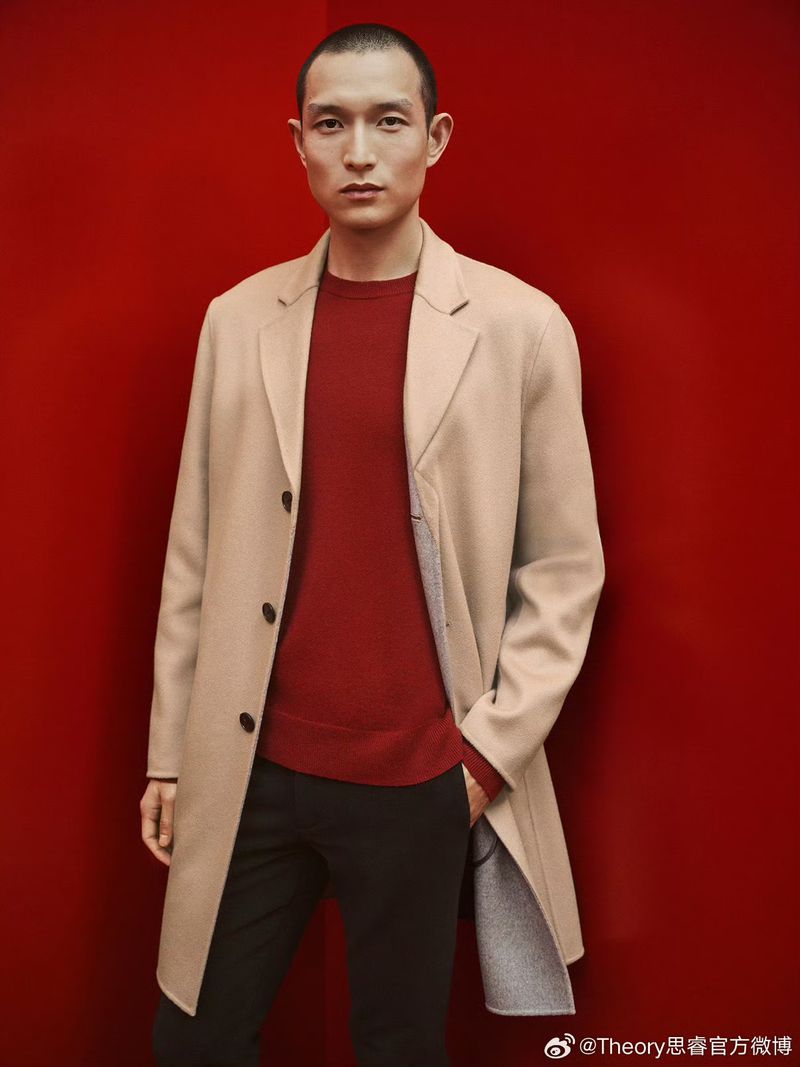 Portrait of an adult male in a beige overcoat and burgundy sweater against a red background in a studio with medium shot.