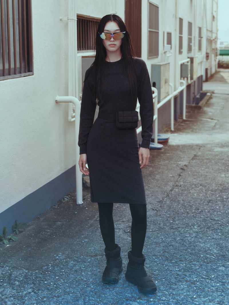 Adult woman in a dark dress, black leggings, and boots stands outside a building with white walls and multiple windows, wearing sunglasses.