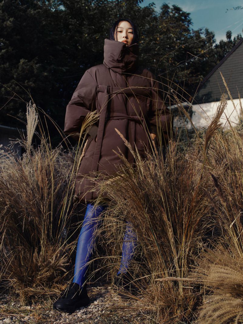 Woman in a brown puffer coat posing in tall grass in front of a house and trees.  She is wearing blue tights and black shoes.