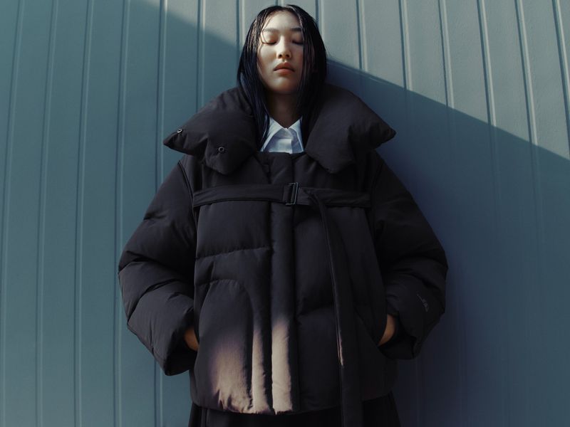 Woman in black puffer jacket posing against a textured blue wall with sunlight, showcasing a contemporary fashion aesthetic.