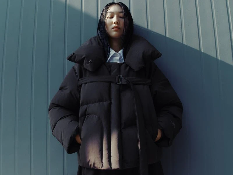 Woman in black puffer jacket posing against a textured blue wall with sunlight, showcasing a contemporary fashion aesthetic.