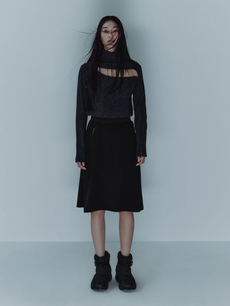 Full-length portrait of an Asian woman wearing a gray long-sleeve cutout top, black skirt, and black puffer boots in a studio.