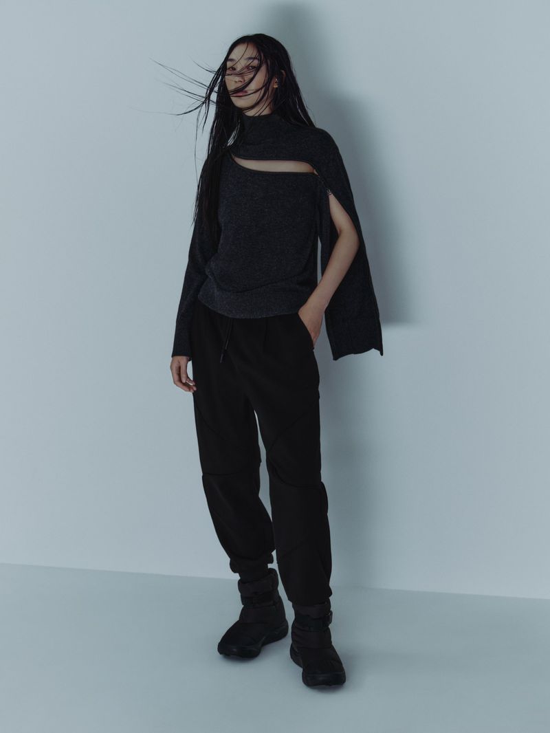 Model with long dark hair blowing in the wind, wearing a cut-out grey sweater and black trousers against a neutral background.