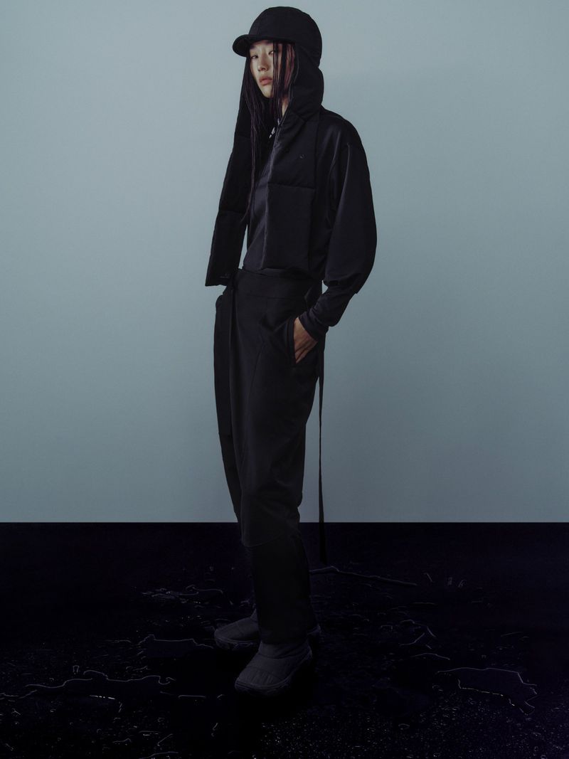 An adult female model poses in a studio wearing a black jacket, top, pants and a cap; full shot against a blue-gray background.