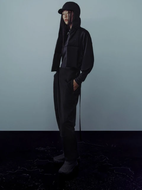 An adult female model poses in a studio wearing a black jacket, top, pants and a cap; full shot against a blue-gray background.