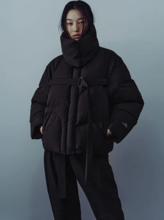 Model wearing a black puffer jacket with a high collar and strapped belt against a muted grey studio backdrop.