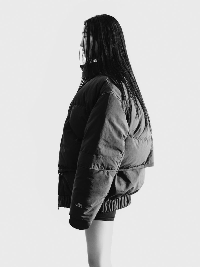 Profile shot of a person with long, dark hair wearing a large puffer jacket against a bright white backdrop in a studio.