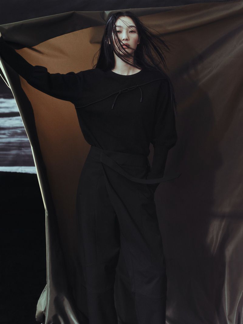 Indoor fashion portrait of an Asian female model wearing a black outfit against a draped background, wind blowing hair and fabric.