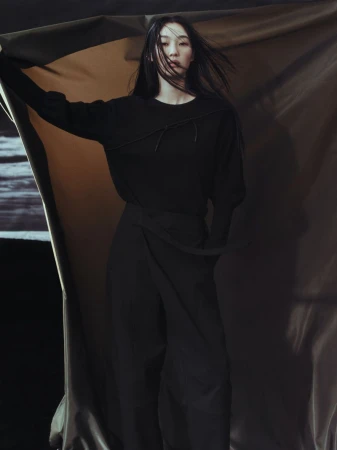 Indoor fashion portrait of an Asian female model wearing a black outfit against a draped background, wind blowing hair and fabric.