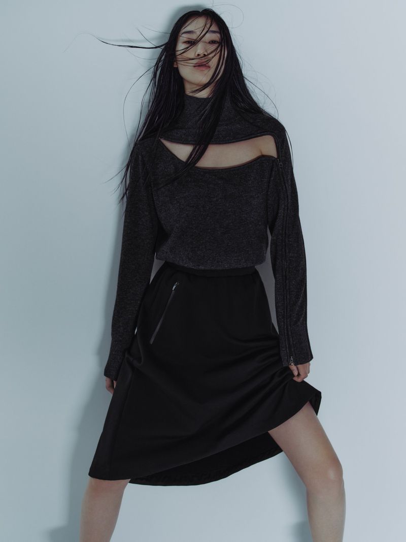 A fashionable woman with long black hair poses in a cutout gray sweater and black skirt against a neutral background, styled for editorial.