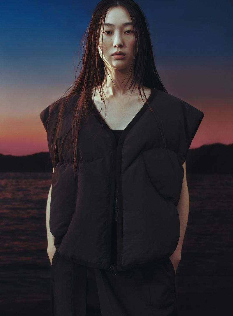 Portrait of Asian woman with long dark hair wearing a black puffer vest in front of a lake at dusk with an orange and blue sky.