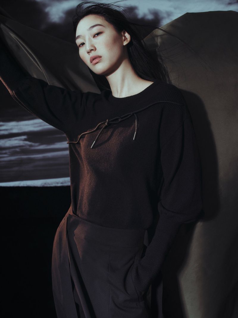 A woman wearing a dark brown sweater and draped skirt poses in a photo studio with a cloudy sky background, illuminated by soft lighting.