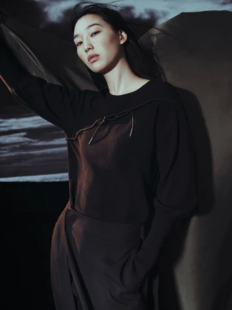 A woman wearing a dark brown sweater and draped skirt poses in a photo studio with a cloudy sky background, illuminated by soft lighting.