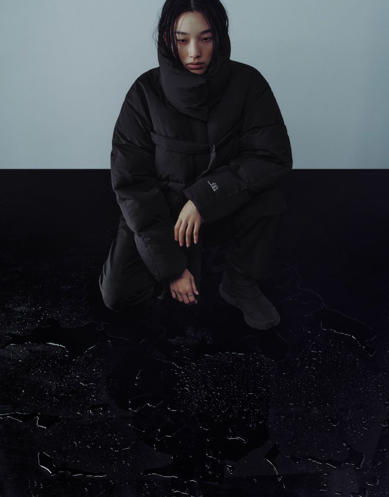 A woman with dark hair in a black puffer jacket kneels on a shiny black surface with water droplets, against a gray and black background.