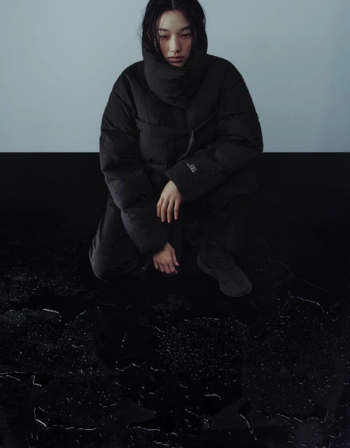 A woman with dark hair in a black puffer jacket kneels on a shiny black surface with water droplets, against a gray and black background.