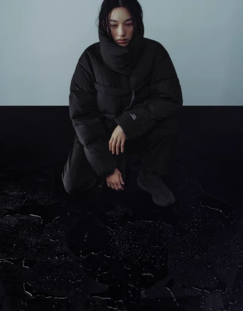 A woman with dark hair in a black puffer jacket kneels on a shiny black surface with water droplets, against a gray and black background.