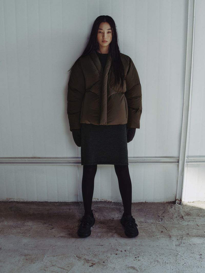 A slender, dark-haired woman models an olive-green puffer jacket with a black dress, tights, and sneakers against a neutral wall.