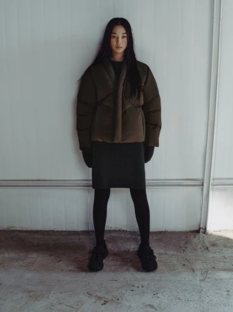 A slender, dark-haired woman models an olive-green puffer jacket with a black dress, tights, and sneakers against a neutral wall.