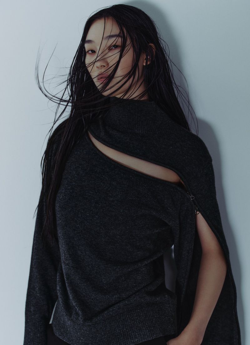 Portrait of an adult female model with long dark hair wearing a dark grey sweater with cut-out design against a light grey backdrop.