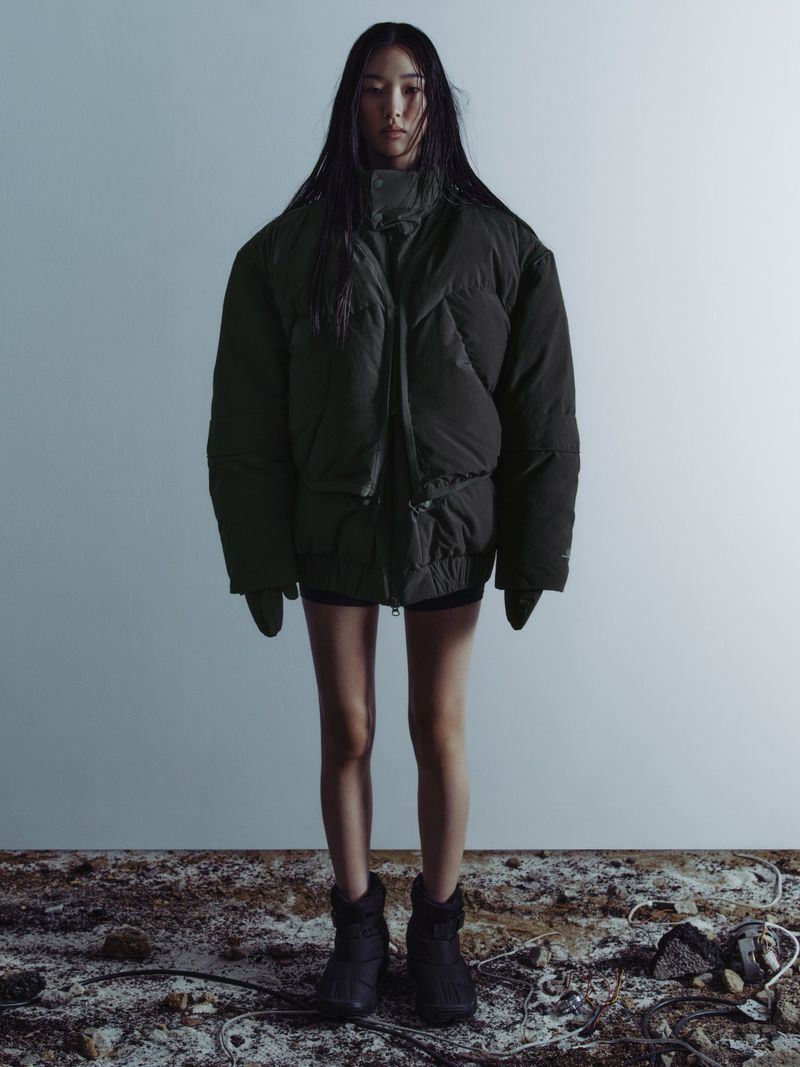 Young woman in olive puffer jacket stands against a neutral background on textured floor. Dark hair, dark boots, cool fashion shot.